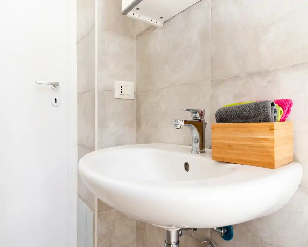 Lavabo de baño moderno y luminoso con grifo cromado, paredes alicatadas y toallas dobladas en un soporte de madera — perfecto para un aseo compacto y limpio.