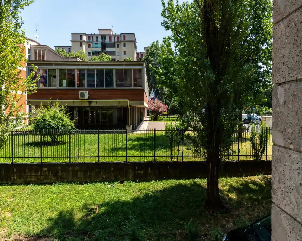Vista de un área exterior vallada con césped, árboles maduros y un edificio bajo moderno al fondo; la luz diurna y la claridad hacen que la imagen sea adecuada para mostrar el espacio exterior.