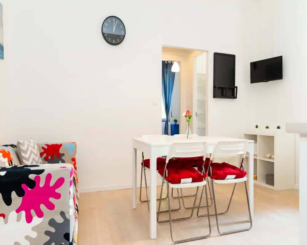 Área de estar luminosa y compacta con mesa blanca, cuatro sillas con cojines rojos y un sofá de estampado colorido: estilo moderno y sencillo, ideal para estancias cortas.