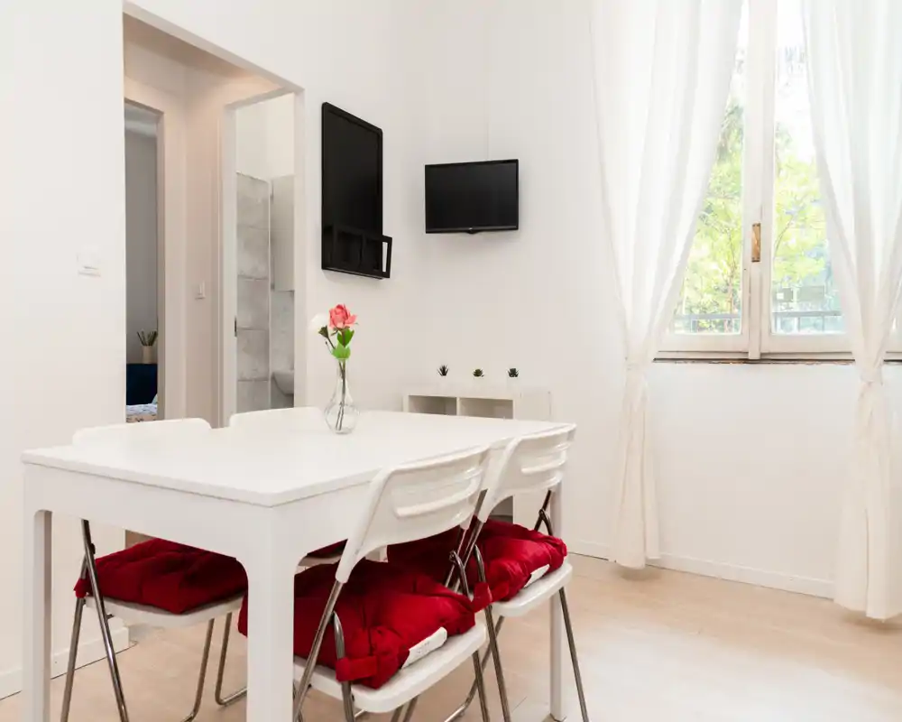 Área de comedor luminosa y minimalista con mesa blanca, cuatro sillas con cojines rojos y un jarrón con flores, bien iluminada por una gran ventana.
