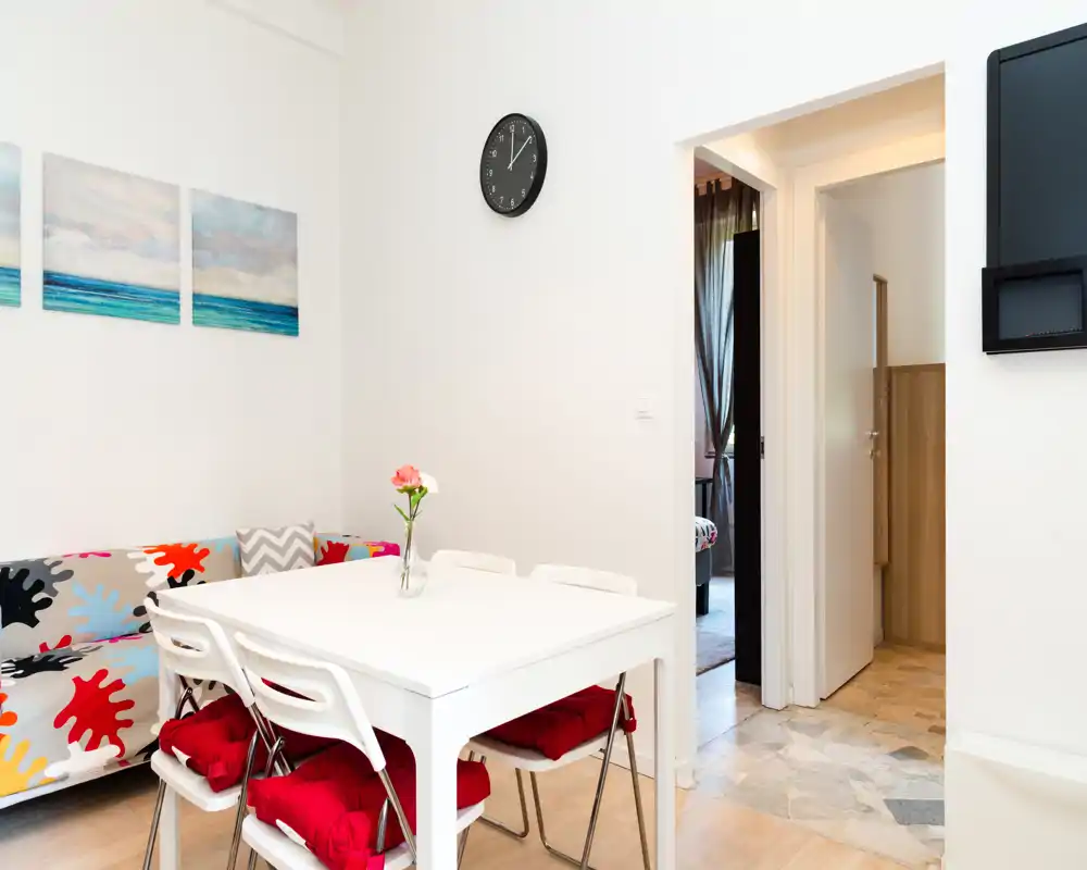 Acogedor comedor pequeño y luminoso con mesa blanca, cuatro sillas con cojines rojos y un pequeño jarrón en el centro; sofá colorido y cuadros en la pared crean un ambiente alegre.