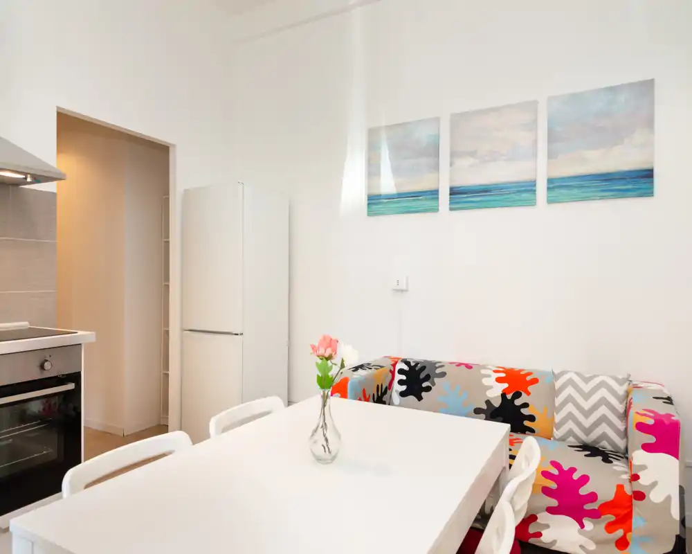 Acogedor comedor de concepto abierto junto a la cocina, con mesa blanca, sillas y un sofá de colores con cuadros en la pared — perfecto para comidas informales.