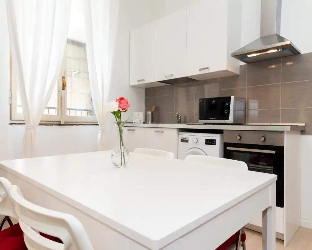 Cocina pequeña moderna y luminosa con mesa de comedor blanca, horno integrado, lavadora y microondas. Un jarrón con una flor aporta calidez; ventana grande con cortinas transparentes que dejan pasar luz natural.