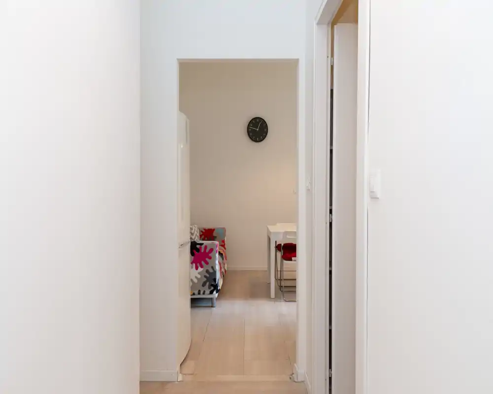 Vista desde el pasillo hacia una pequeña sala con un sofá colorido y un reloj de pared; paredes neutras y decoración minimalista.