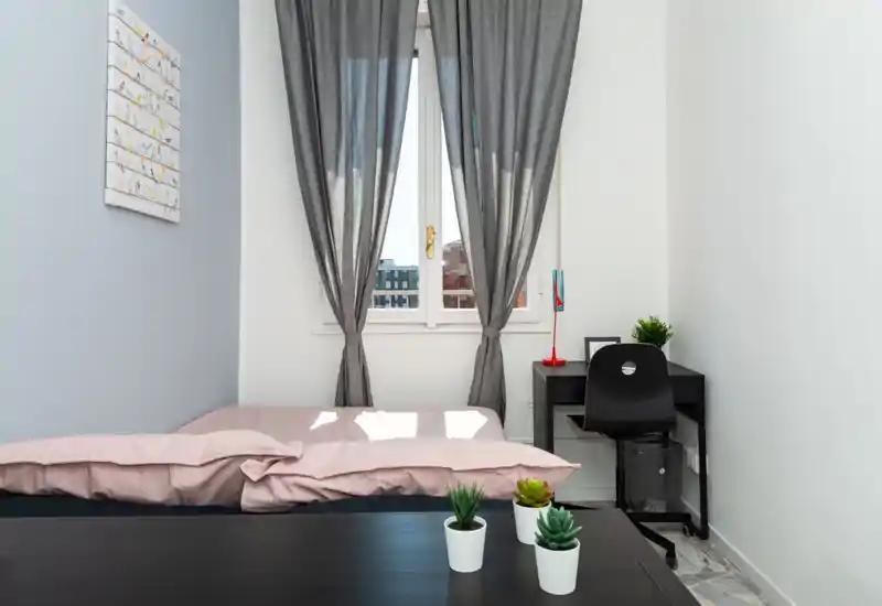 Dormitorio individual luminoso y bien decorado con edredón rosa, ventana central con cortinas grises y un escritorio compacto — ideal para una estancia acogedora.