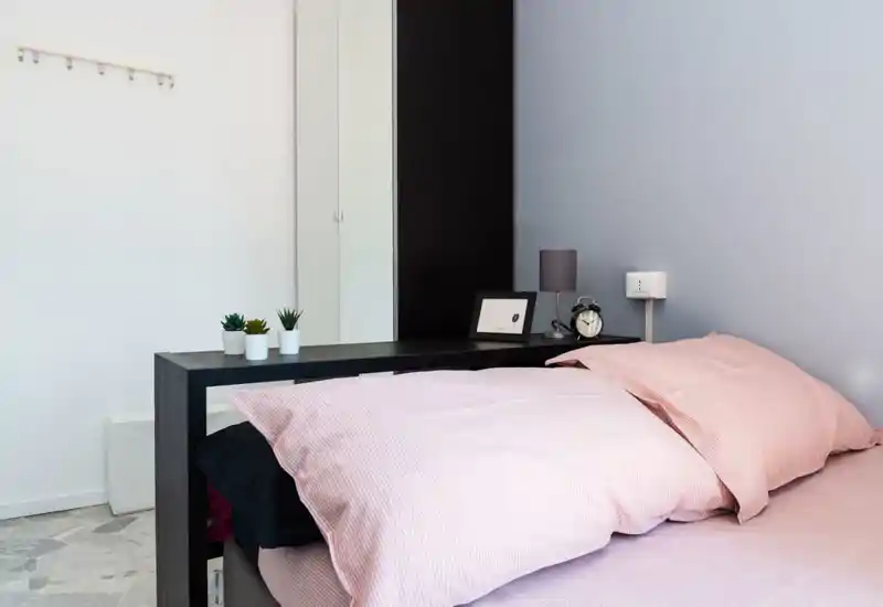 Dormitorio moderno y luminoso con cama vestida en sábanas rosa, mesita con lámpara y despertador, y pequeñas plantas decorativas que crean un ambiente tranquilo y acogedor.