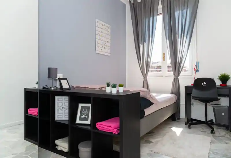 Dormitorio individual luminoso y ordenado con cama bien hecha junto a la ventana. La habitación cuenta con una estantería negra al pie de la cama, un escritorio y silla, textiles suaves y plantas que aportan calidez.