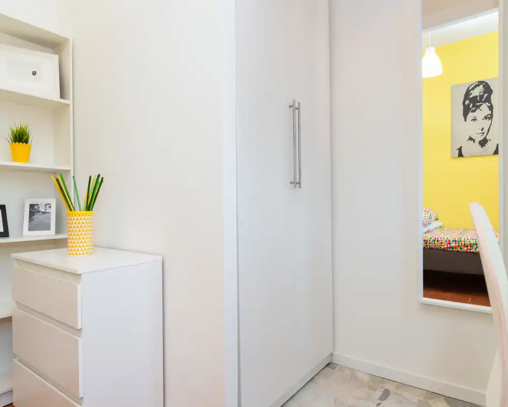 Small, bright bedroom corner with white storage and a wardrobe; a mirror reflects a colorful bed in an adjacent sleeping area.