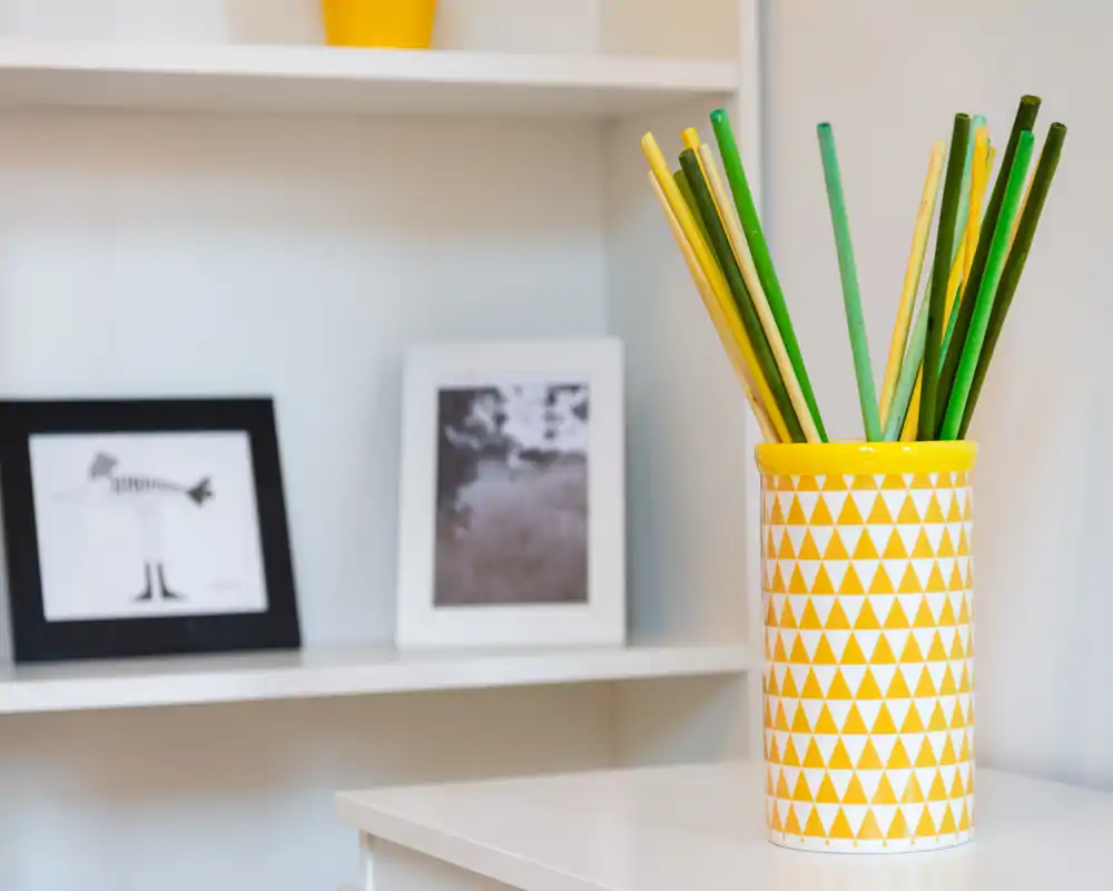Close-up of a bright, modern living area detail: a patterned yellow vase with decorative sticks on a white surface and framed photos on shelving in the background.