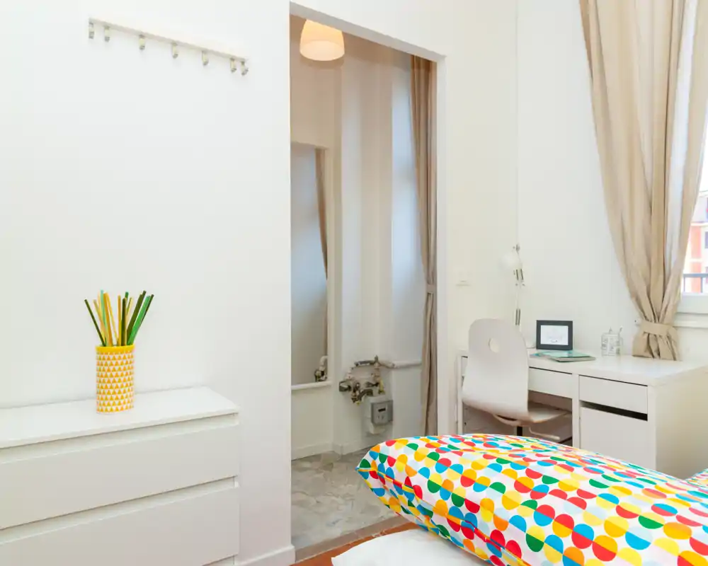 Bright, modern bedroom with colorful bedding in the foreground, a small desk by the window and clean white furnishings. Natural light and tidy styling create a welcoming atmosphere ideal for rental photos.