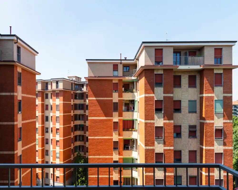Vista desde un balcón hacia edificios residenciales cercanos con una barandilla metálica en primer plano; imagen diurna nítida que muestra fachadas de ladrillo rojo y ventanas.