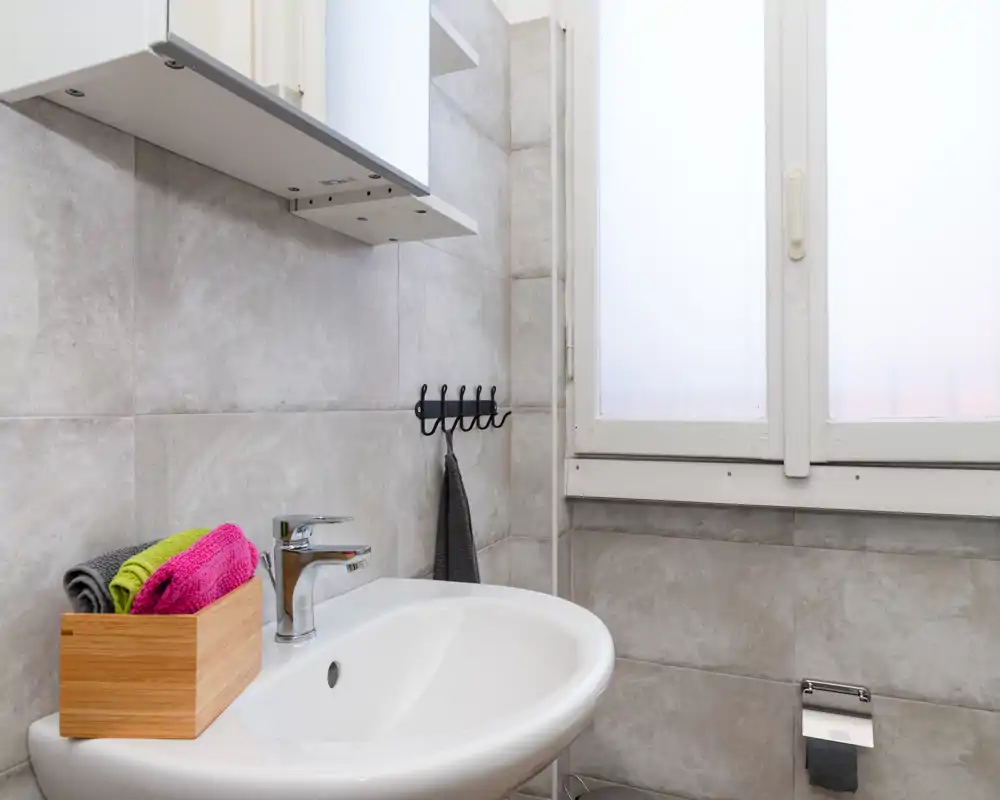 Pequeña zona de baño moderna con lavabo, grifo cromado, azulejos en la pared, ganchos para toallas y ventana traslúcida que aporta privacidad y luz suave.