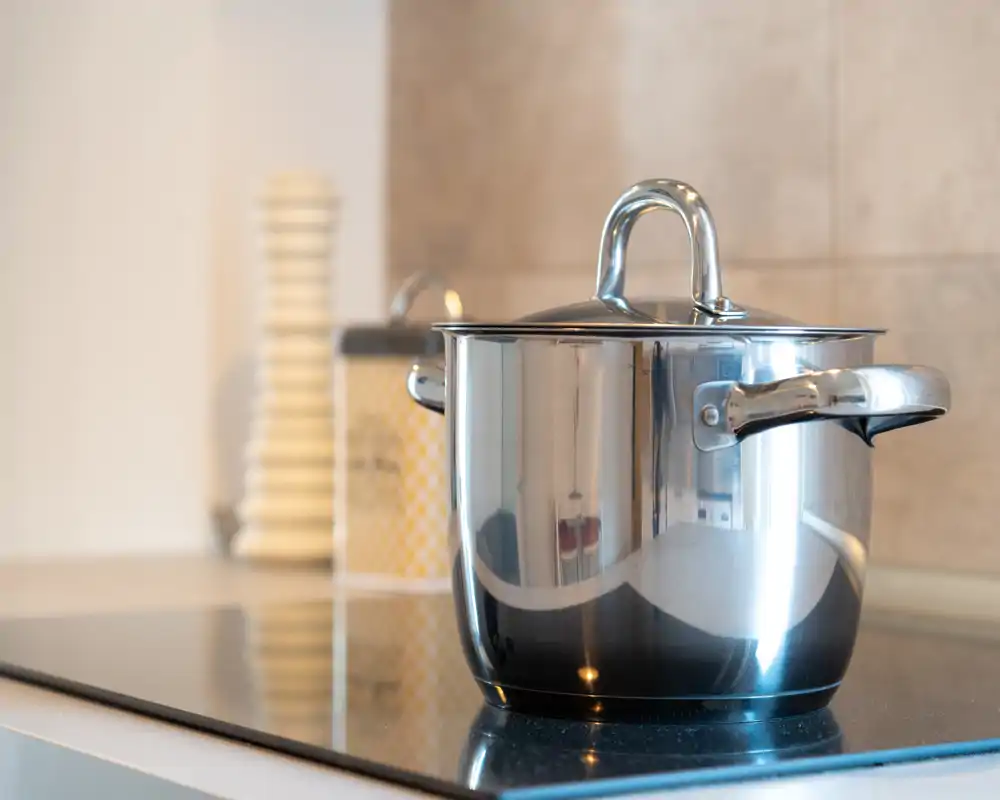 Plano cercano de una cacerola de acero inoxidable sobre una placa de inducción moderna; superficie de cocina limpia y bien iluminada con tarros decorativos al fondo.