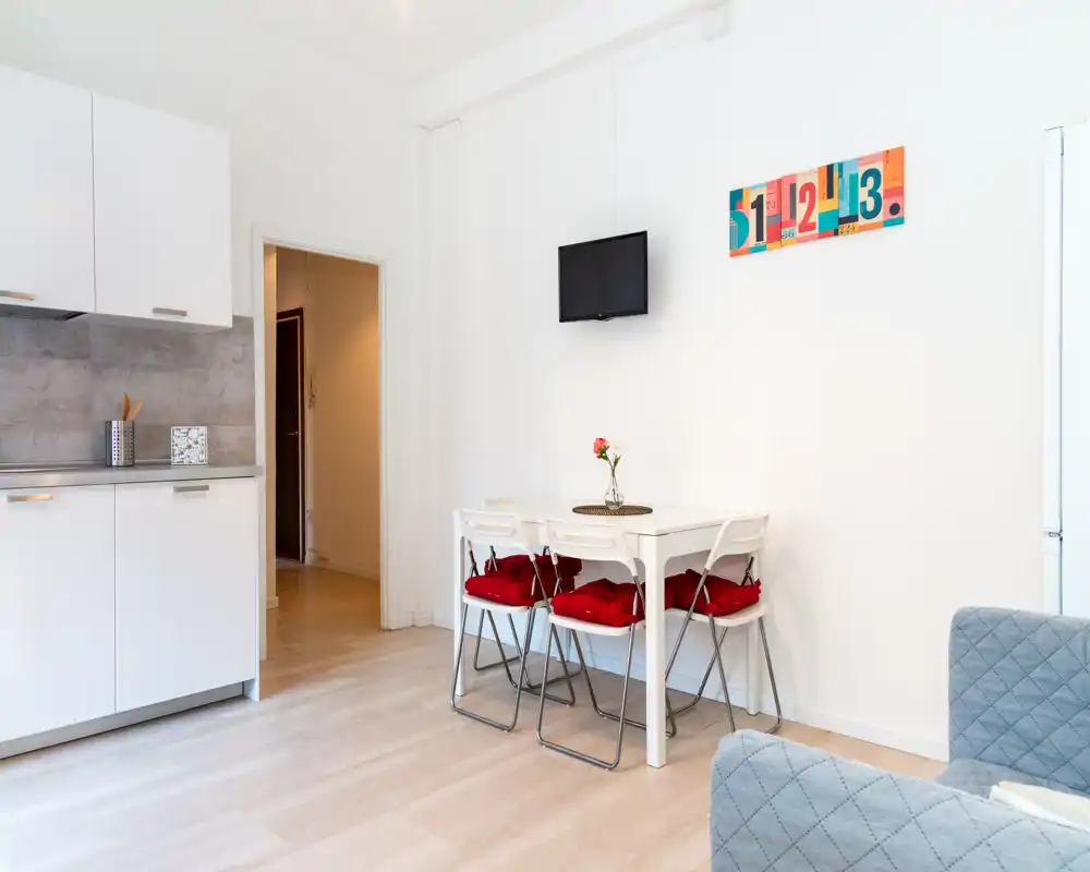 Cocina abierta y zona de comedor compacta con muebles blancos, mesa pequeña con cuatro sillas con cojines rojos y televisor en la pared — luminosa, limpia y moderna, ideal para estancias cortas.