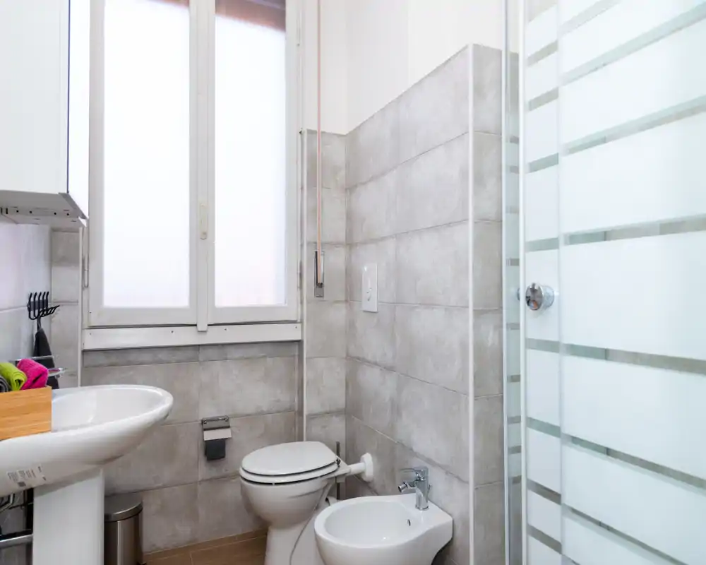 Baño compacto y moderno con paredes alicatadas, lavabo de pedestal, inodoro, bidé y puerta de ducha de vidrio esmerilado. La luz natural de una ventana alta aporta una sensación limpia y funcional.