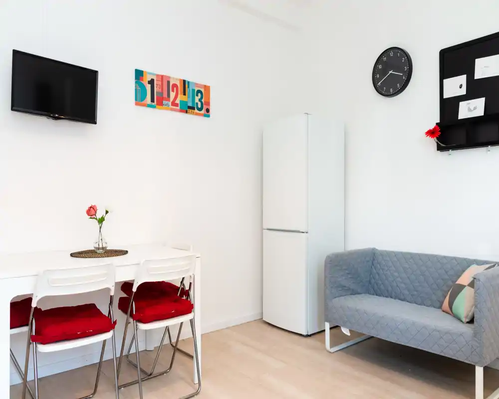 Zona de estar luminosa y compacta con un sofá gris de dos plazas moderno, mesa de comedor con sillas con cojines rojos y frigorífico junto a la pared — estilo limpio y minimalista, ideal para un apartamento pequeño.