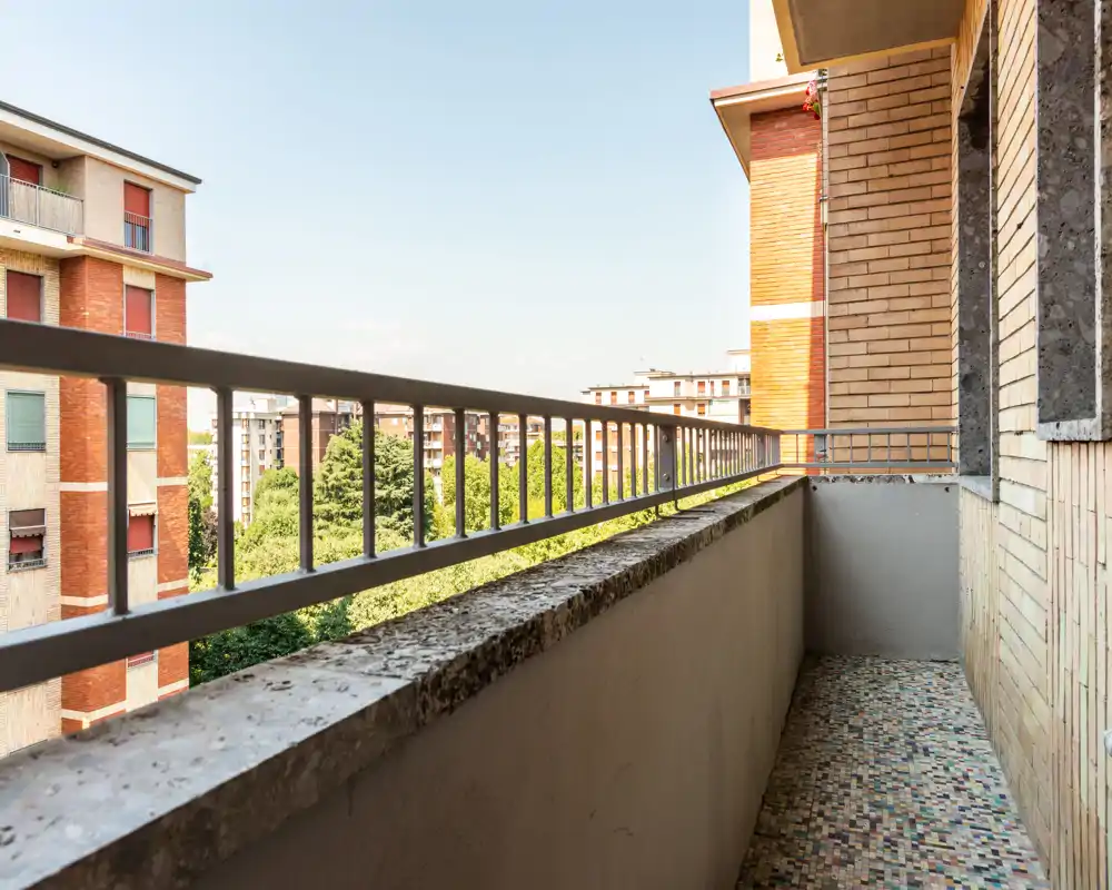 Balcón estrecho y soleado con suelo de baldosas y barandilla metálica, con vistas a árboles y edificios vecinos — ideal para una pequeña zona de descanso o plantas.