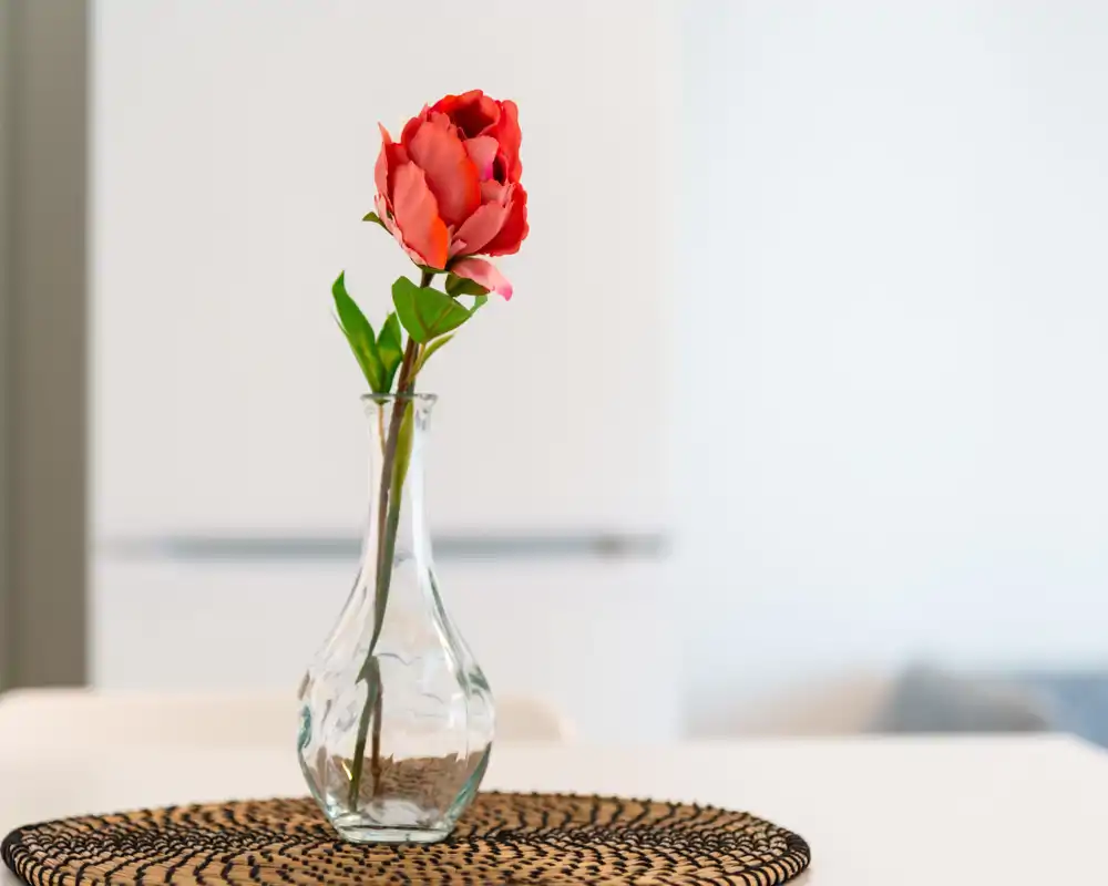 Primer plano de una flor roja en un jarrón de cristal sobre un salvamantel tejido, fotografiada en una mesa luminosa. El estilo minimalista sugiere un rincón de comedor o desayuno.