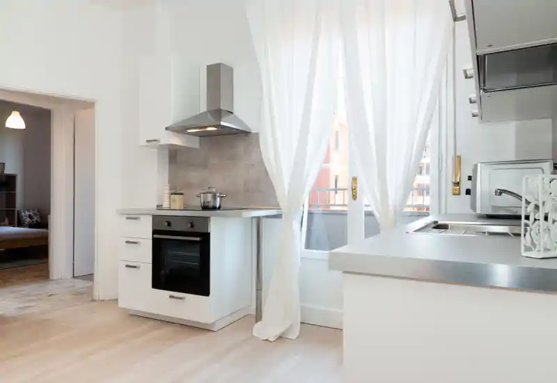 Modern, compact kitchen with white cabinetry, built-in oven, stainless hood and bright natural light from a window with soft curtains.