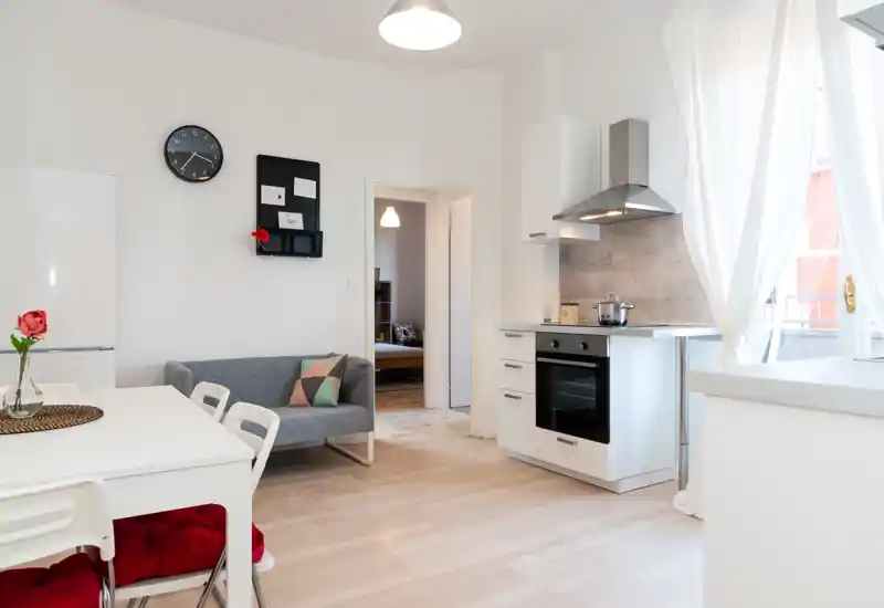 Bright, modern kitchen area with integrated oven, extractor hood and a small dining table adjacent to a cozy seating nook. Large window with white curtains fills the space with natural light.