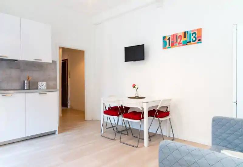Small modern kitchen corner with white cabinets, a compact dining table with four chairs and red cushions, and a mounted TV — bright, clean and tastefully decorated for a cozy stay.