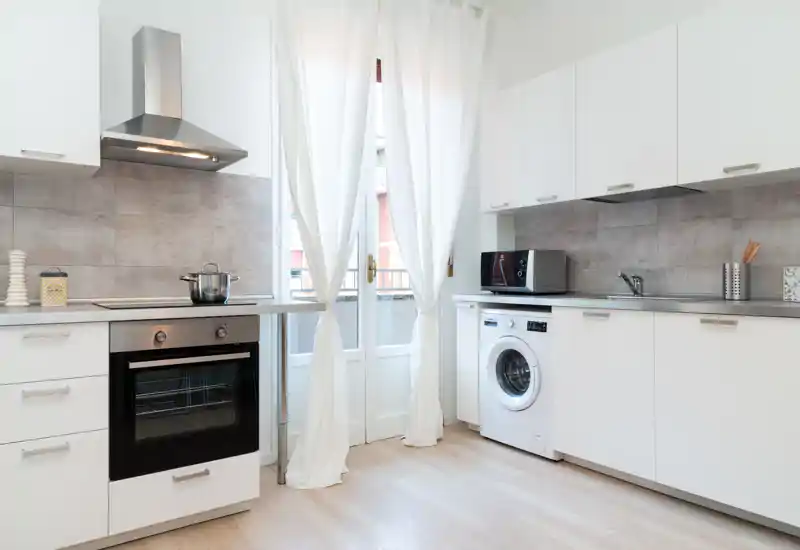 Modern, bright kitchen with white cabinetry, built-in oven, induction hob, and a washing machine; sheer curtains soften the natural light and create a fresh, inviting cooking space.