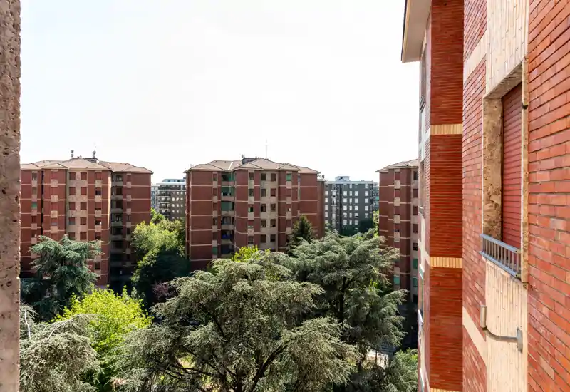 Bright balcony view overlooking mature trees and neighboring red-brick apartment buildings — ideal to showcase the outdoor space and urban vista.