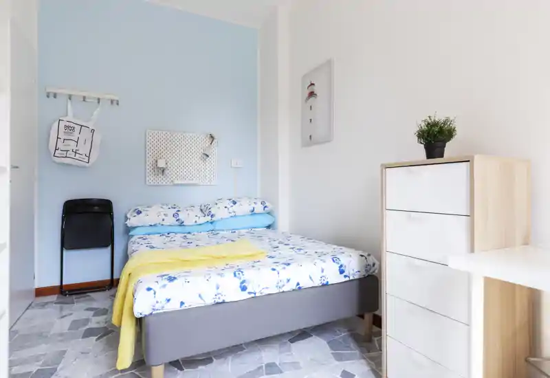 Sunny, well-staged bedroom with a double bed dressed in floral linens, a pale blue feature wall and a compact chest of drawers — bright and welcoming for short-term rentals.