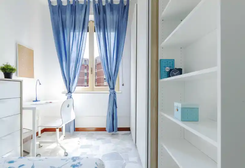 Bright, compact bedroom with blue curtains, a small desk and shelving — ideal for a single occupant or student. Natural light fills the space and the clean, minimal styling makes it feel fresh and calm.