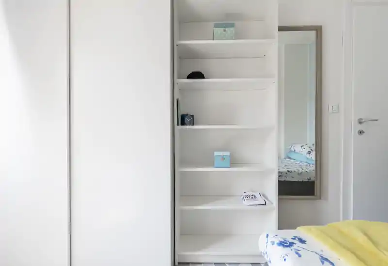 Bright, minimal bedroom corner with a partial view of the bed, built-in shelving and a full-length mirror. The clean white palette and floral textiles create a fresh, uncluttered sleeping area.