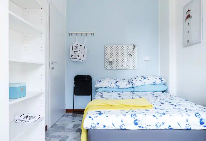 Bright, compact bedroom with a double bed dressed in blue floral linens, soft pillows and a yellow throw. Clean, modern styling with light-blue walls and practical shelving nearby — ideal for a cozy short-stay.