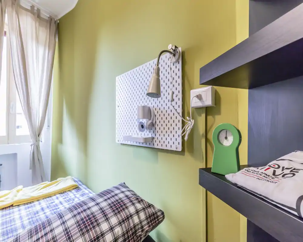 Small bedroom with a patterned single bed beside a bright window; soft natural light and a wall-mounted reading lamp create a cozy, well-styled sleeping corner.