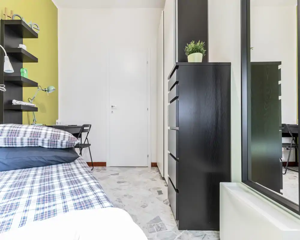 Bright, modern bedroom with a partially visible double bed dressed in checkered linens, a small desk and shelving on the left, and a tall black dresser with a mirrored wardrobe on the right.