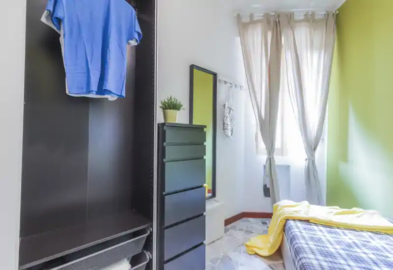 Bright single bedroom with a neatly made bed, patterned bedding and a tall black dresser; natural light filters through sheer curtains creating a fresh, airy atmosphere.