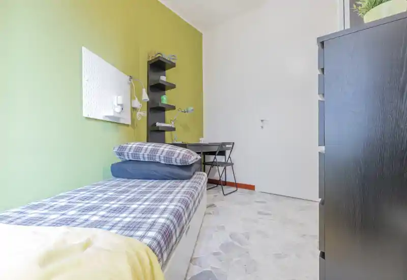 Bright, tidy single bedroom with a neatly made bed, plaid bedding and a compact desk area. Green accent wall and shelving create a fresh, modern feel ideal for a student or short-term rental.