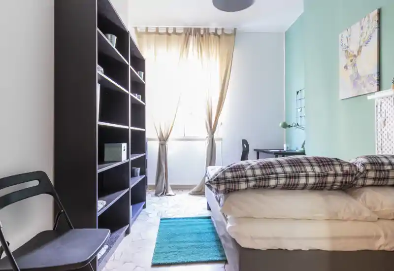 Dormitorio luminoso y ordenado con cama bien hecha, pared de acento turquesa y estantería junto a la ventana soleada — acogedor y bien iluminado.