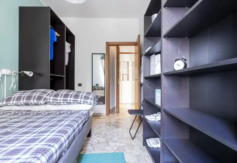 Dormitorio luminoso y bien compuesto con cama doble y ropa de cama a cuadros, estanterías abiertas y un pequeño armario; suelo de baldosas limpio y un espejo que aporta profundidad.