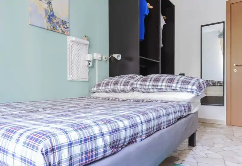 Dormitorio luminoso y ordenado con una cama doble con ropa de cama de cuadros, lámpara de lectura y armario abierto. Suelo de baldosas y espejo de cuerpo entero completan este espacio compacto y bien iluminado.