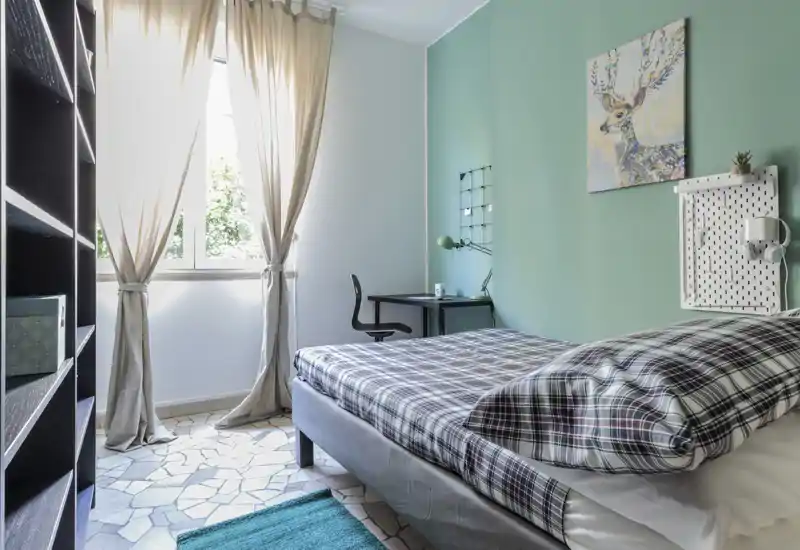 Dormitorio luminoso y acogedor con ropa de cama a cuadros, un escritorio junto a la ventana y luz natural suave. Pared en verde, cuadro decorativo y cama doble ordenada.