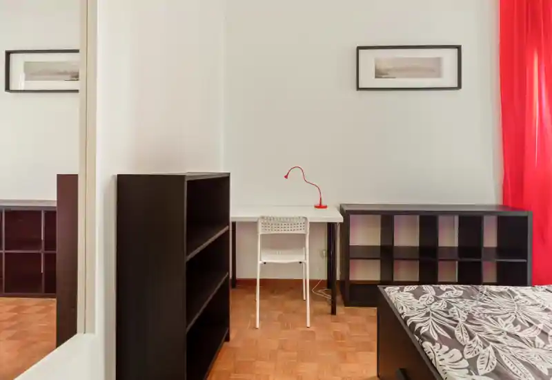 Bright, simply furnished bedroom corner with a single bed partially visible, a desk with a red lamp and modular storage units; clean parquet flooring and a bold red curtain add character.
