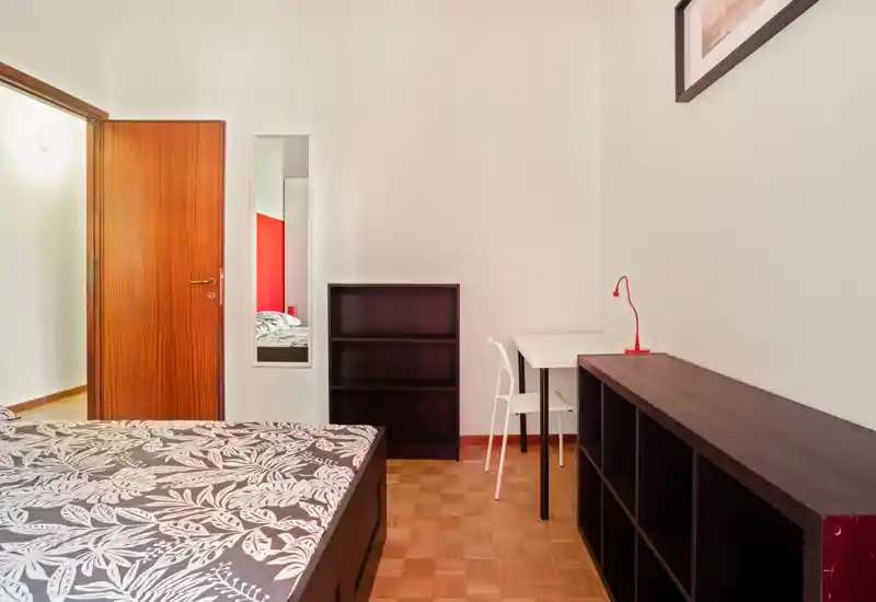 Bright, tidy bedroom with a patterned double bed in the foreground, dark wood shelving and a small desk with a red lamp — ideal for short-term rentals or student stays.