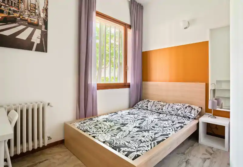 Bright, neatly styled bedroom with a wooden double bed, patterned bedding, bedside table with lamp and a warm orange accent wall; natural light enters through a curtained window.