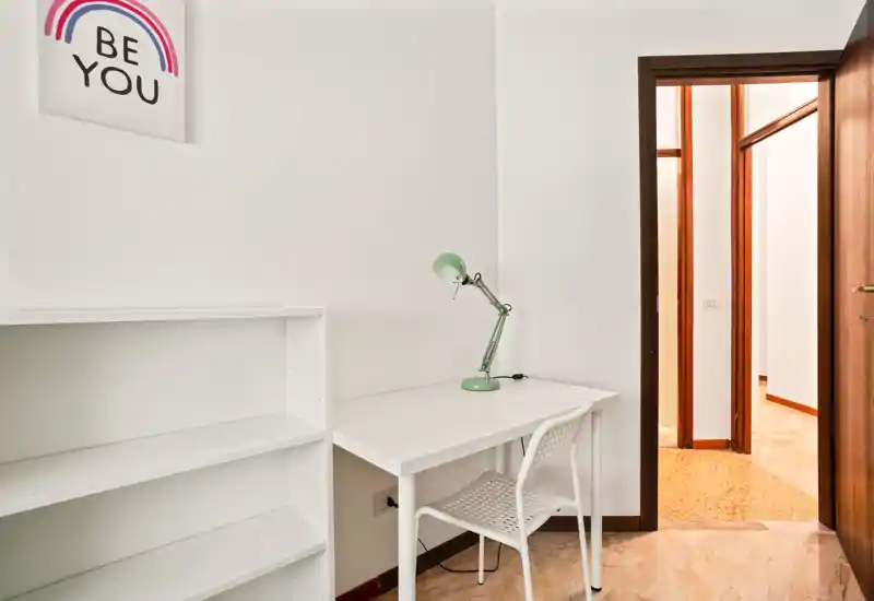 Small, minimal entrance/study nook with a white desk, shelving and a green desk lamp; bright neutral walls and a doorway leading to the hallway.