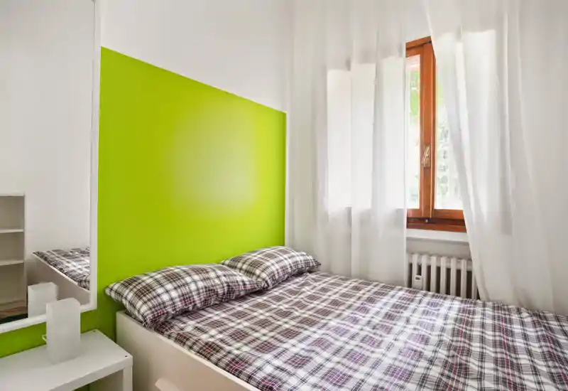 Bright, compact bedroom with a double bed against a vivid green accent wall, plaid bedding and a large window providing natural light.