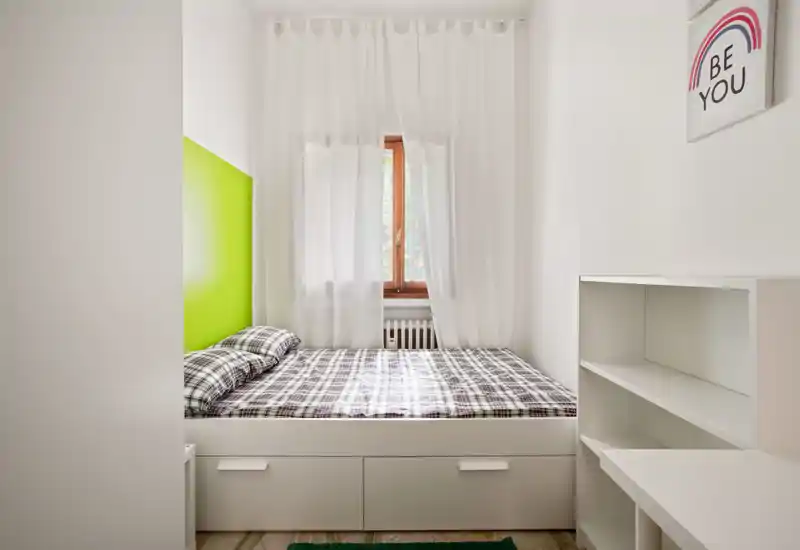 Bright, compact bedroom with a full bed framed by built-in storage drawers, a green accent headboard and sheer curtains letting in natural light.