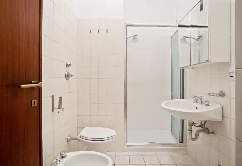 Clean, compact bathroom with a wall-mounted sink, bidet and toilet, plus a tiled walk-in shower with glass door. Neutral white tiles and good lighting create a bright, functional space ideal for short-term rentals.