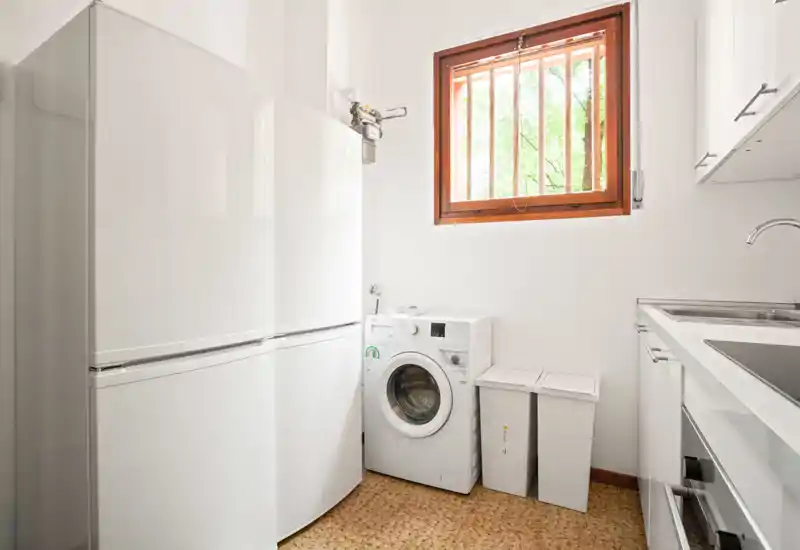 Compact, bright kitchen/laundry corner with a fridge, washing machine and sink; clean white finishes and natural light from a wooden-framed window.