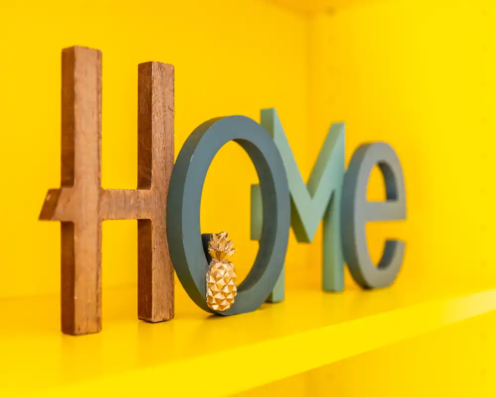 Primer plano de letras decorativas de madera y pintadas que forman “HOME” sobre una estantería amarilla brillante; detalle decorativo sin contexto de la habitación.