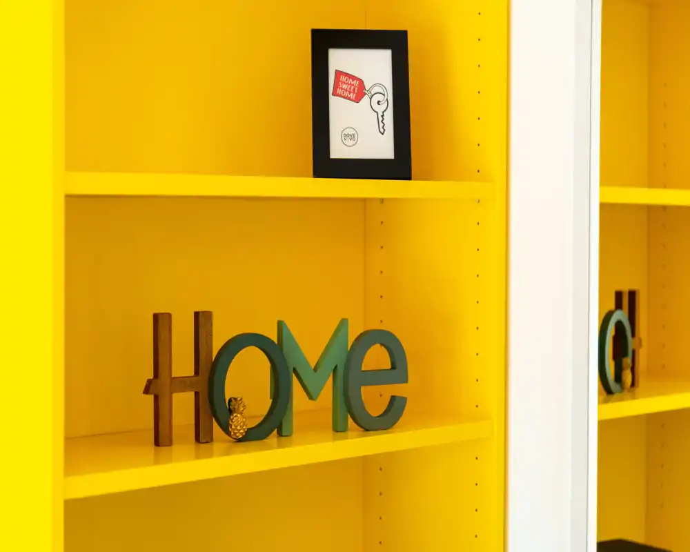 Primer plano de estantería amarilla brillante con objetos decorativos que forman “HOME”; la imagen se centra en la estantería y la decoración, no en una habitación concreta.