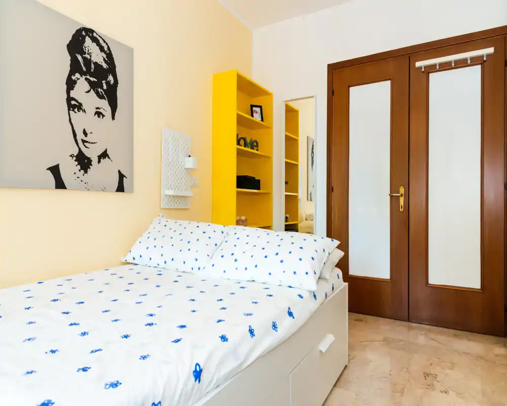 Dormitorio luminoso y bien presentado con cama doble con ropa de cama blanca con motivos azules, estantería amarilla y cuadro decorativo; espacio de descanso limpio y acogedor.